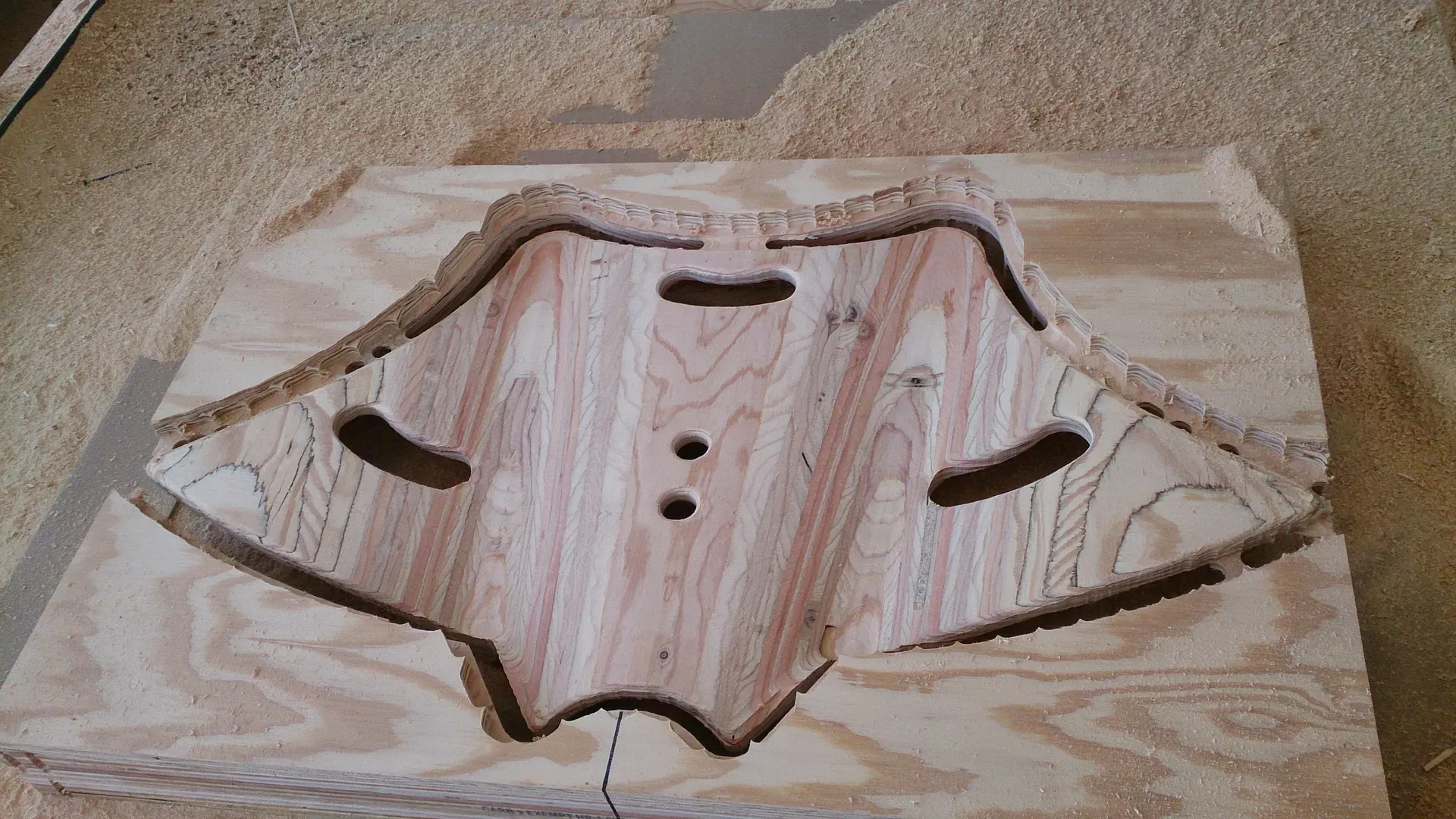 A complex, organic-shaped component freshly cut from a sheet of plywood using our CNC router. The piece features precisely routed curves and internal cutouts, still seated within the original board. This highlights our precision manufacturing capabilities and the raw, clean finish of our woodworking process.