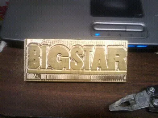 A custom-machined brass stamping die featuring the "BIGSTAR" logo resting on a workbench next to a laptop and pliers.