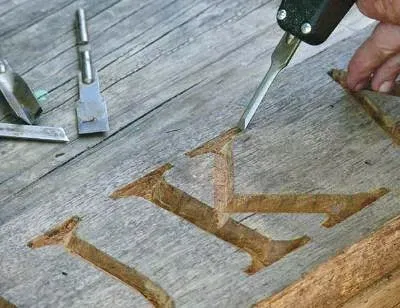 A close-up view of a wood chisel carving the letter 'K' into a piece of weathered, reclaimed timber, showcasing traditional hand-carving techniques in a woodworking studio.