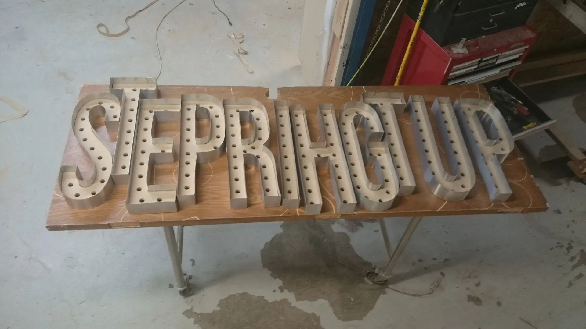 This image shows custom-fabricated metal channel letters spelling out "STEP UP," laid out on a workshop table during the assembly phase. The galvanized steel letter returns have been shaped and pre-drilled to house future lighting components. These letters are currently being prepared for wiring and face installation, representing the high-quality craftsmanship and structural integrity of our custom signage production process.